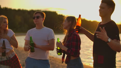 The-teens-spend-the-carefree-summertime-at-sunset-on-the-sand-beach-with-beer.-They-are-talking-to-each-other-and-and-laugh-at-funny-stories-on-the-open-air-party.
